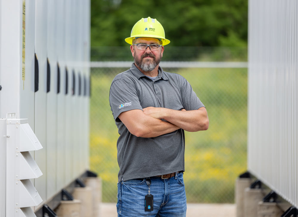An Alliant Energy employee in a yellow hard hat stands with his arms crossed in front of white battery cabinets.